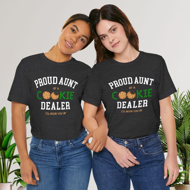 May include: Two women wearing dark gray t-shirts with the text "PROUD AUNT OF A COOKIE DEALER" in white and green, with a cookie graphic. The phrase "I'LL HOOK YOU UP" is below the text. They are wearing blue jeans.