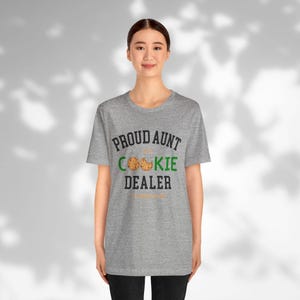 May include: A heather gray t-shirt with the text "PROUD AUNT OF COOKIE DEALER" in black and green. The word "COOKIE" features a cookie graphic. The shirt is worn by a person.