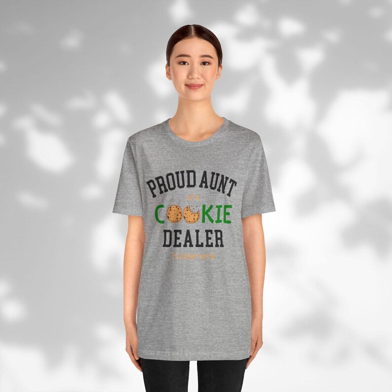 May include: A heather gray t-shirt with the text "PROUD AUNT OF COOKIE DEALER" in black and green. The word "COOKIE" features a cookie graphic. The shirt is worn by a person.