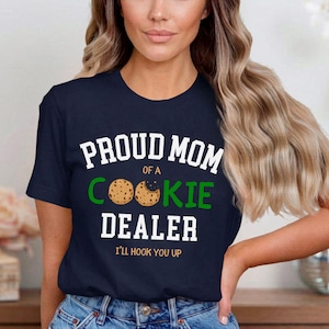 May include: Navy blue t-shirt with the text "PROUD MOM of a COOKIE DEALER" in white and green. The design includes cookie graphics. The phrase "I'LL HOOK YOU UP" is printed below the main text.