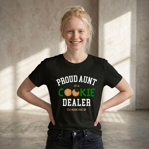 May include: Black t-shirt with the text "PROUD AUNT of a COOKIE DEALER I'LL HOOK YOU UP" in white and green. The shirt features a cookie graphic. The person is smiling and wearing dark jeans.