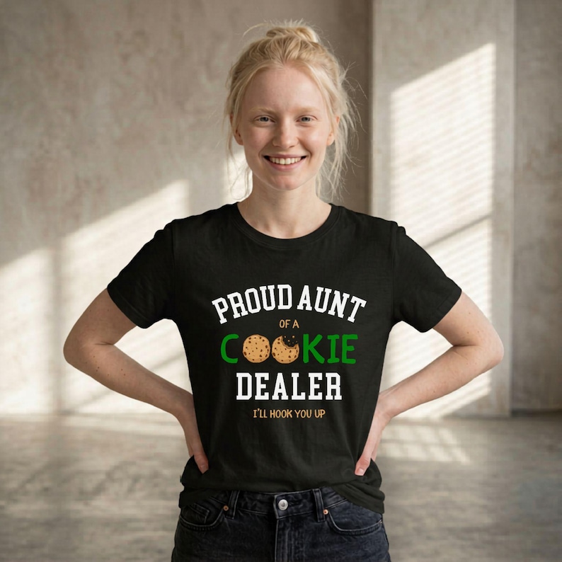 May include: Black t-shirt with the text "PROUD AUNT of a COOKIE DEALER I'LL HOOK YOU UP" in white and green. The shirt features a cookie graphic. The person is smiling and wearing dark jeans.