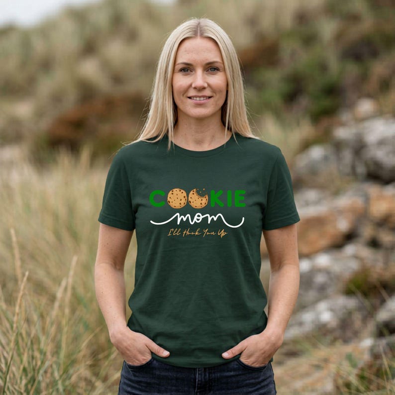 May include: A dark green t-shirt with the words "COOKIE mom" in green and white script. The word "COOKIE" has two cookie graphics. Below the words is the phrase "I'll Hook You Up". The shirt is being worn by a person with blonde hair.