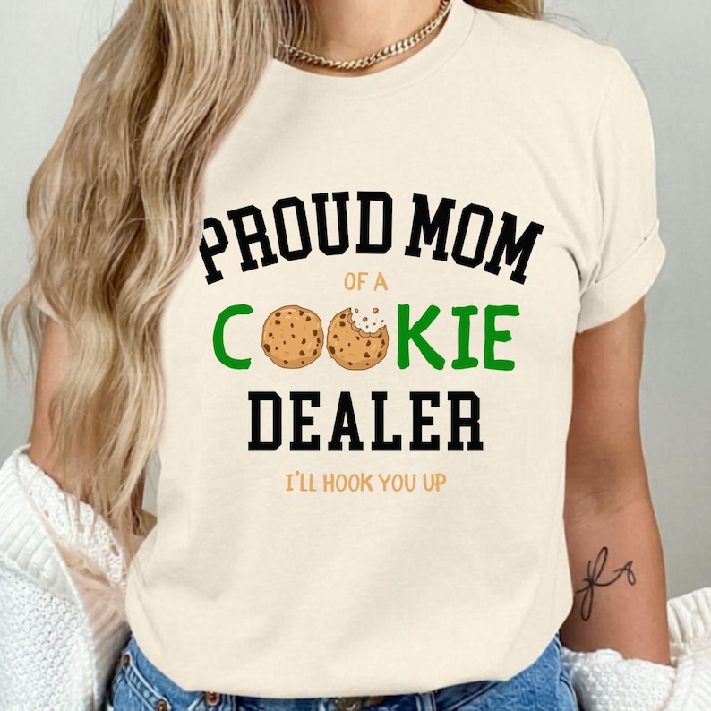 May include: Cream-colored t-shirt with the text "PROUD MOM OF A COOKIE DEALER" in black and green. The word "COOKIE" features two cookie illustrations. Below the text is "I'LL HOOK YOU UP" in black.