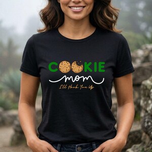 May include: Black t-shirt with the words "COOKIE mom" in green and white. The word "COOKIE" has two cookie images as the letters "O" and "O". Below the word "mom" is the phrase "I'll Hook You Up".