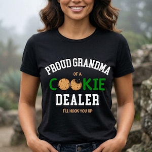 May include: Black t-shirt with the text "PROUD GRANDMA OF A COOKIE DEALER" in white and green. The word "COOKIE" features a cookie graphic. The phrase "I'LL HOOK YOU UP" is printed below the main text.