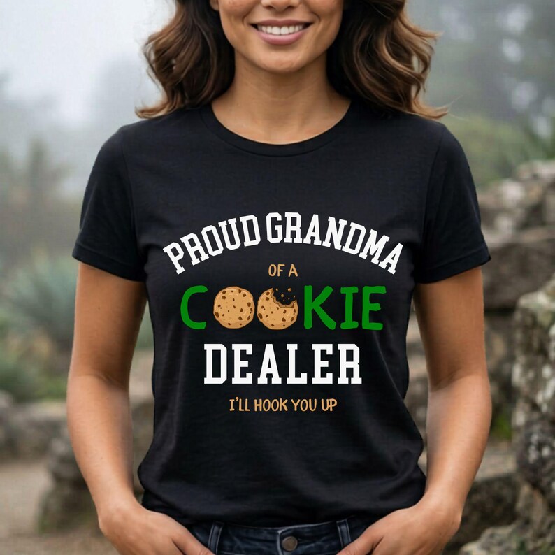 May include: Black t-shirt with the text "PROUD GRANDMA OF A COOKIE DEALER" in white and green. The word "COOKIE" features a cookie graphic. The phrase "I'LL HOOK YOU UP" is printed below the main text.
