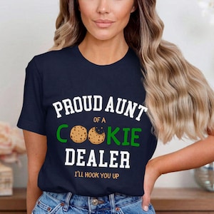 May include: Navy blue t-shirt with the text "PROUD AUNT OF A COOKIE DEALER" in white and green. The design includes cookie graphics and the phrase "I'LL HOOK YOU UP" in white. The shirt is a casual wear item.