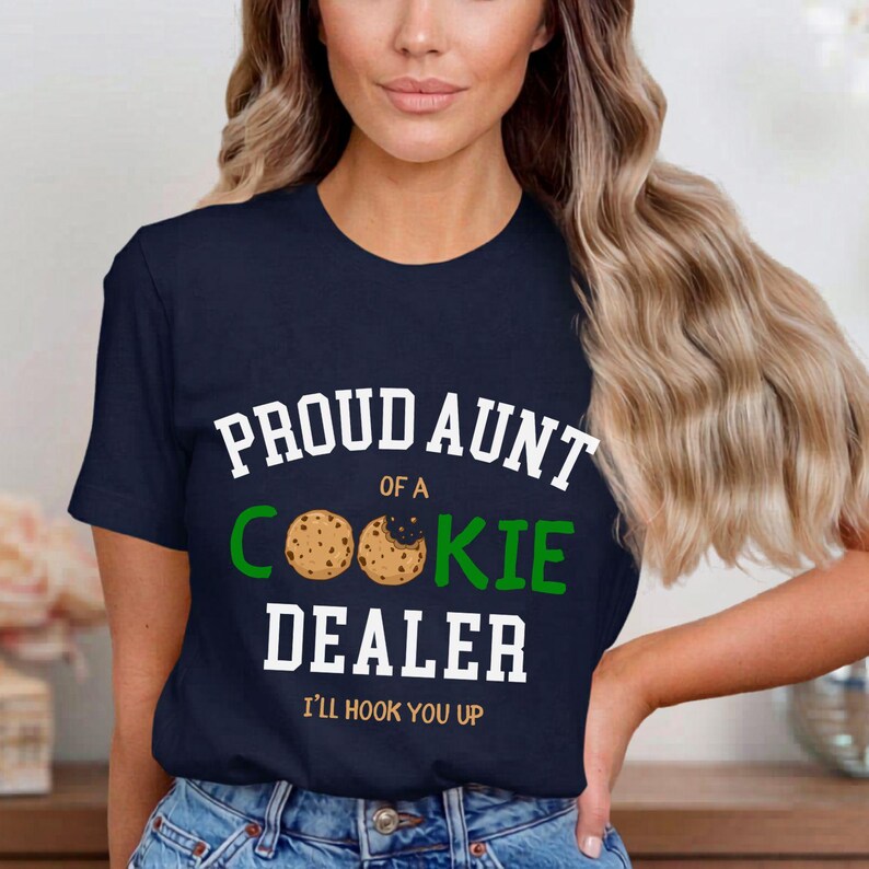 May include: Navy blue t-shirt with the text "PROUD AUNT OF A COOKIE DEALER" in white and green. The design includes cookie graphics and the phrase "I'LL HOOK YOU UP" in white. The shirt is a casual wear item.