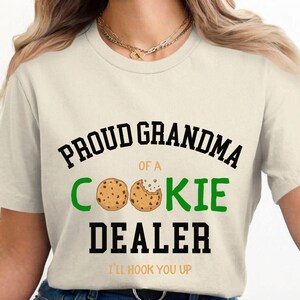 May include: Cream-colored t-shirt with the text "PROUD GRANDMA OF A COOKIE DEALER" in black and green. Two cookie illustrations are featured. The phrase "I'LL HOOK YOU UP" is printed at the bottom.
