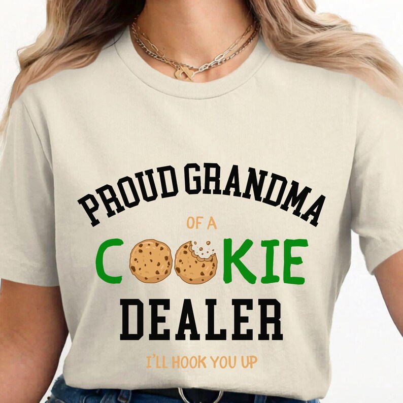 May include: Cream-colored t-shirt with the text "PROUD GRANDMA OF A COOKIE DEALER" in black and green. Two cookie illustrations are featured. The phrase "I'LL HOOK YOU UP" is printed at the bottom.