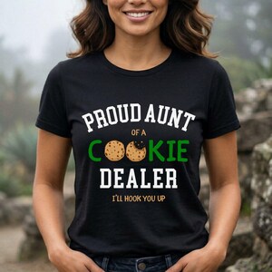May include: Black t-shirt with the text "PROUD AUNT OF A COOKIE DEALER" in white and green. The word "COOKIE" features two cookie graphics. The phrase "I'LL HOOK YOU UP" is printed below the main text.