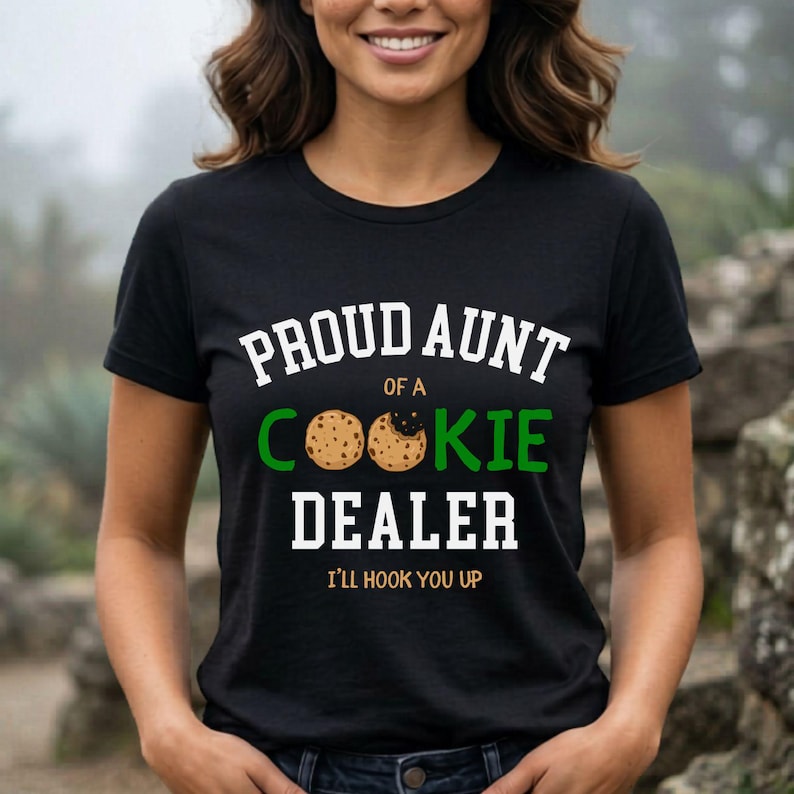 May include: Black t-shirt with the text "PROUD AUNT OF A COOKIE DEALER" in white and green. The word "COOKIE" features two cookie graphics. The phrase "I'LL HOOK YOU UP" is printed below the main text.