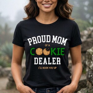 May include: Black t-shirt with the text "PROUD MOM of a COOKIE DEALER" in white and green. The word "COOKIE" features a cookie graphic. The phrase "I'LL HOOK YOU UP" is printed below the text.