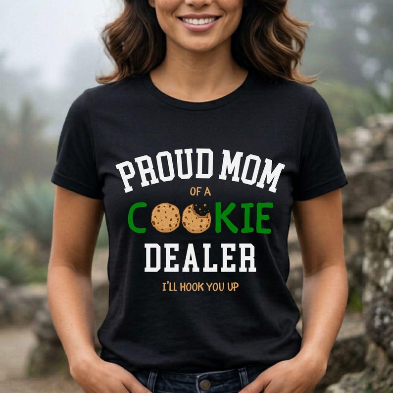 May include: Black t-shirt with the text "PROUD MOM of a COOKIE DEALER" in white and green. The word "COOKIE" features a cookie graphic. The phrase "I'LL HOOK YOU UP" is printed below the text.