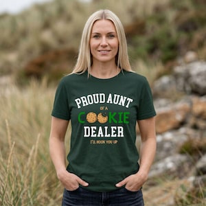 May include: A dark green t-shirt with the text "PROUD AUNT of a COOKIE DEALER" in white and green. The shirt features a cookie graphic and the phrase "I'LL HOOK YOU UP" in a playful font. The woman is smiling.