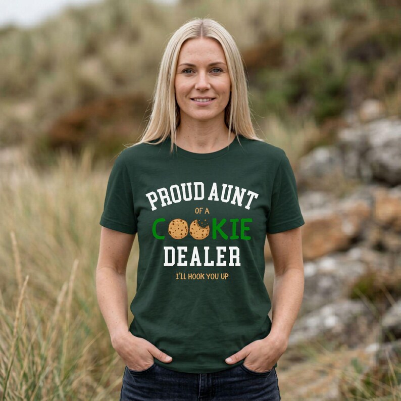 May include: A dark green t-shirt with the text "PROUD AUNT of a COOKIE DEALER" in white and green. The shirt features a cookie graphic and the phrase "I'LL HOOK YOU UP" in a playful font. The woman is smiling.