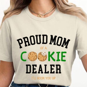 May include: Cream-colored t-shirt with the text "PROUD MOM of a COOKIE DEALER" in black and green. Two cookie illustrations are featured. The phrase "I'LL HOOK YOU UP" is printed at the bottom in orange.
