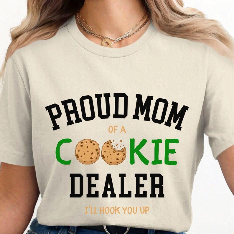 May include: Cream-colored t-shirt with the text "PROUD MOM of a COOKIE DEALER" in black and green. Two cookie illustrations are featured. The phrase "I'LL HOOK YOU UP" is printed at the bottom in orange.