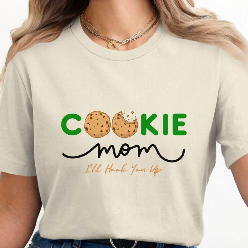 May include: Cream-colored t-shirt with the words "COOKIE mom" in green and black. The word "COOKIE" features two cookie illustrations. Below the words is the phrase "I'll Hook You Up" in a cursive font.