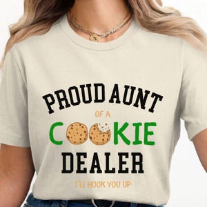 May include: Cream-colored t-shirt with the text "PROUD AUNT of a COOKIE DEALER" in black and green. The word "COOKIE" features two cookie illustrations. Below the text is "I'LL HOOK YOU UP" in orange.