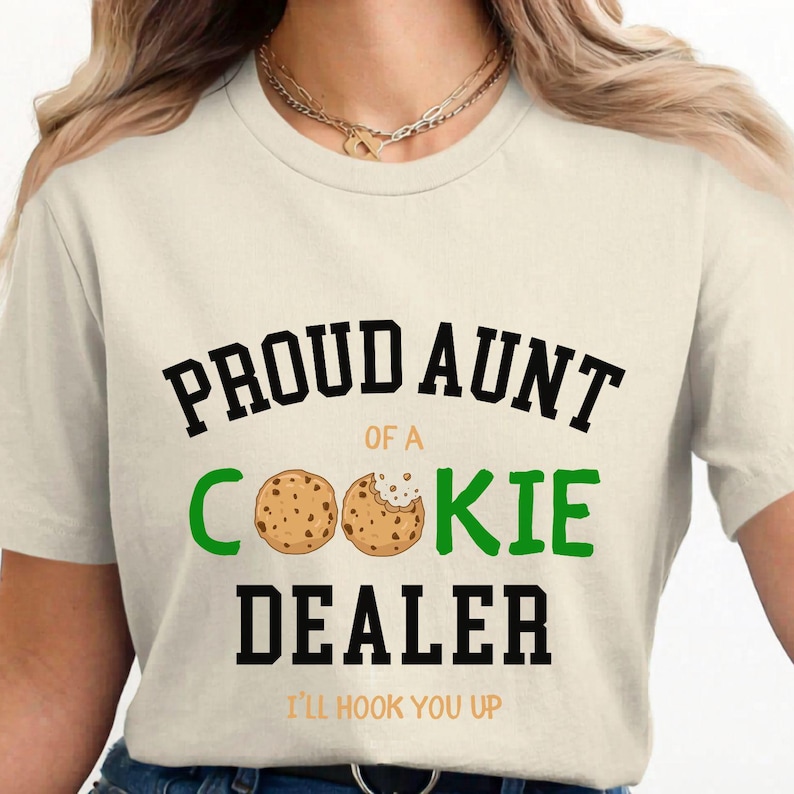 May include: Cream-colored t-shirt with the text "PROUD AUNT of a COOKIE DEALER" in black and green. The word "COOKIE" features two cookie illustrations. Below the text is "I'LL HOOK YOU UP" in orange.