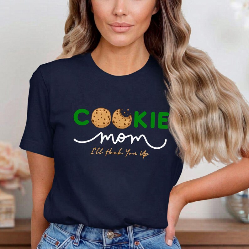 May include: Navy blue t-shirt with the words "COOKIE mom" in green and white, featuring cookie graphics. The text "I'll Hook You Up" is below the word "mom". The shirt is made of a soft material.