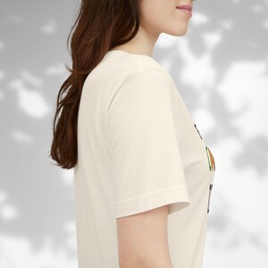 May include: Cream-colored short-sleeve t-shirt with a graphic design. The design includes black, brown, and green elements. The shirt is made of a soft, comfortable material.