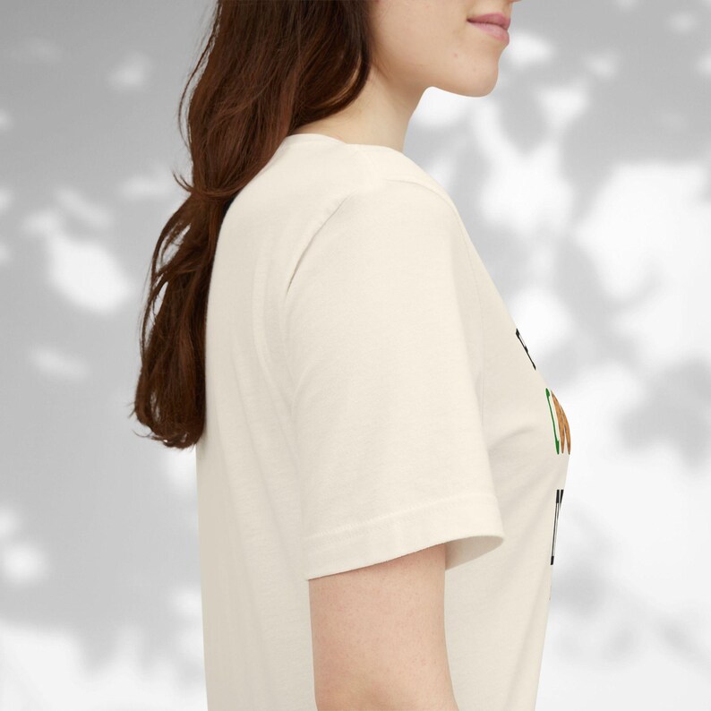 May include: Cream-colored short-sleeve t-shirt with a graphic design. The design includes black, brown, and green elements. The shirt is made of a soft, comfortable material.