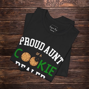 May include: Black t-shirt with the text "PROUD AUNT OF A COOKIE DEALER" in white and green. The design includes two cookie illustrations. The shirt is folded on a dark wood surface.