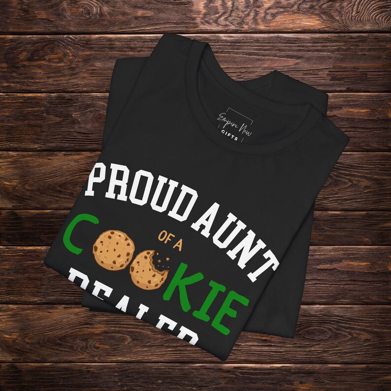 May include: Black t-shirt with the text "PROUD AUNT OF A COOKIE DEALER" in white and green. The design includes two cookie illustrations. The shirt is folded on a dark wood surface.