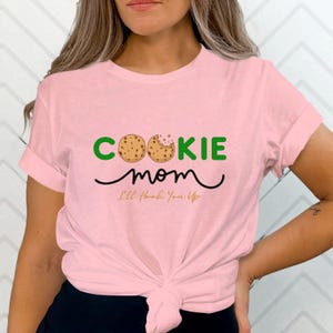 May include: A light pink t-shirt with the words "COOKIE MOM" in green and brown, with a cookie graphic. Below the words is the word "mom" in a cursive font, and the phrase "I'll Hook You Up" in a smaller font.