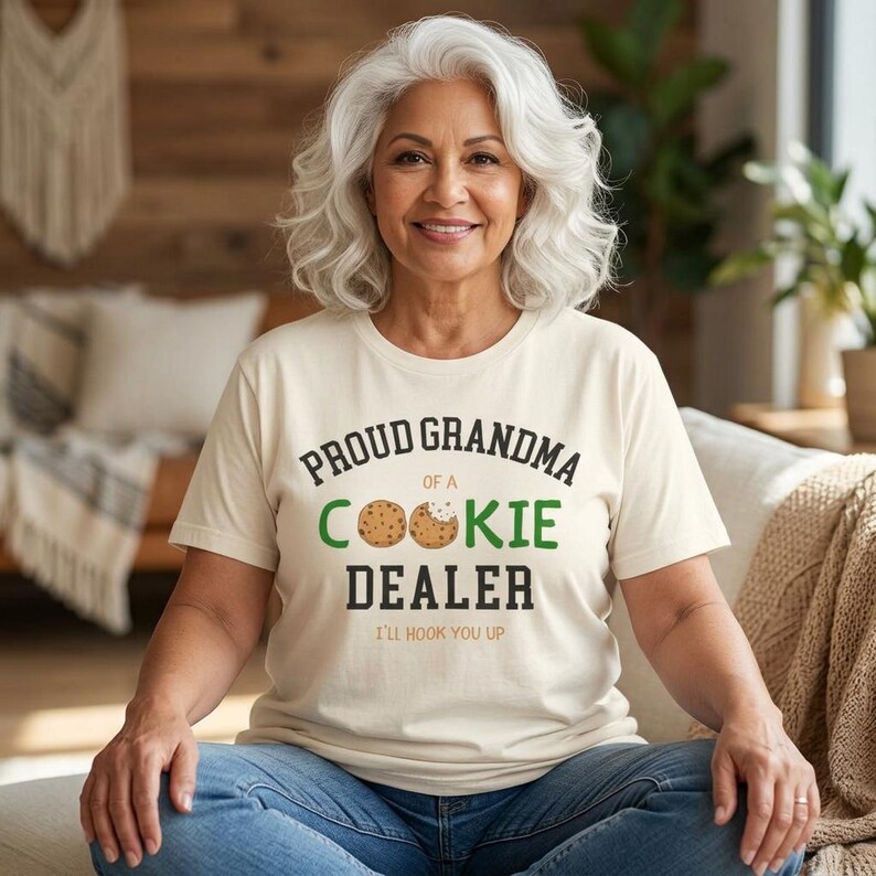 May include: A woman wearing a cream-colored t-shirt with the text "PROUD GRANDMA OF A COOKIE DEALER" in black and green. The shirt features a cookie graphic and the phrase "I'LL HOOK YOU UP". The woman is smiling.