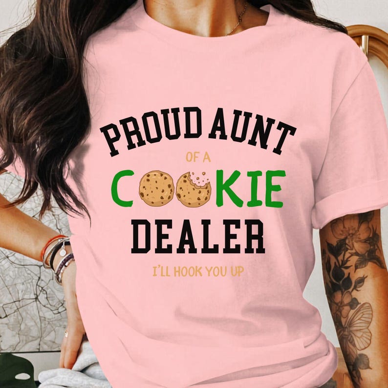 May include: A light pink t-shirt with the text "PROUD AUNT OF A COOKIE DEALER" in black and green. The word "COOKIE" features two cookie illustrations. Below the text is "I'LL HOOK YOU UP" in a smaller font.