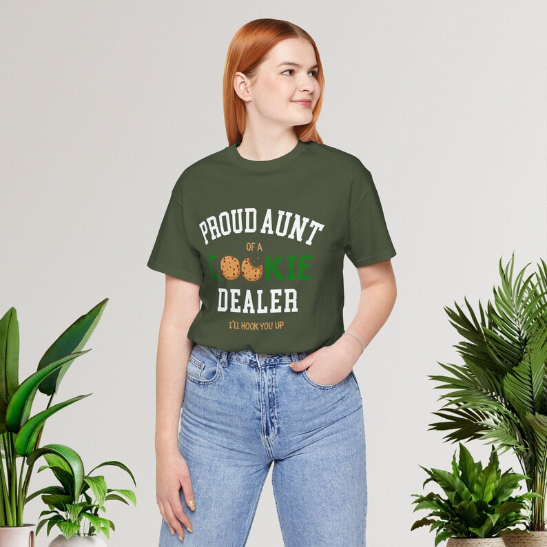 May include: An olive green t-shirt with the text "PROUD AUNT OF A COOKIE DEALER I'LL HOOK YOU UP" in white and green. The design includes cookie graphics. The shirt is worn with blue jeans.