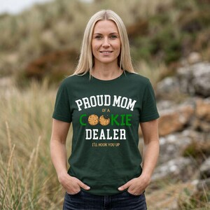 May include: Dark green t-shirt with the text "PROUD MOM of a COOKIE DEALER" in white and green. The shirt features a cookie graphic and the phrase "I'LL HOOK YOU UP" in a playful font. The shirt is a casual wear item.
