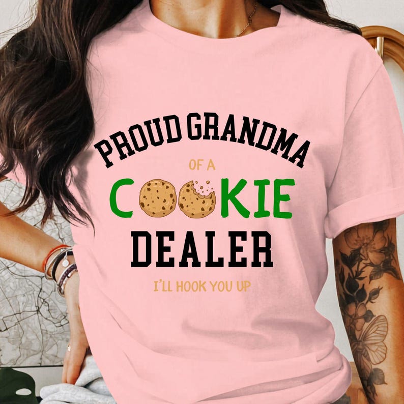 May include: A light pink t-shirt with the text "PROUD GRANDMA of a COOKIE DEALER" in black and green. Two cookie illustrations are between the words "COOKIE" and "DEALER". The phrase "I'LL HOOK YOU UP" is at the bottom.