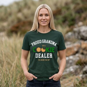 May include: A dark green t-shirt with the text "PROUD GRANDMA of a COOKIE DEALER" in white and green. The shirt features a cookie graphic and the phrase "I'LL HOOK YOU UP" in white. The woman is smiling.