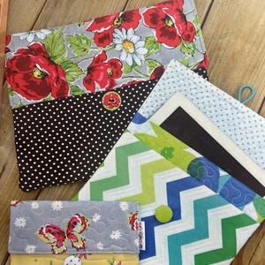May include: Three fabric pouches with different patterns and colors. The top pouch has a floral pattern with red flowers and green leaves on a gray background. The middle pouch has a black and white polka dot pattern. The bottom pouch has a blue, green, and yellow chevron pattern. Each pouch has a button closure.