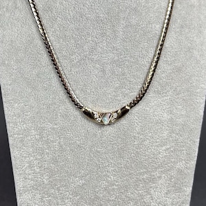 Puede incluir: Un collar de cadena en tono dorado con una textura tejida y trenzada en todos los eslabones. El centro del collar muestra un pequeño elemento colgante triangular acentuado con una piedra central iridiscente y dos pequeñas piedras claras a cada lado.