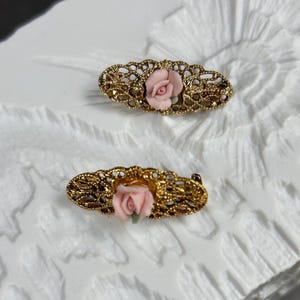 May include: Two vintage gold-tone brooches. Each brooch has an oval filigree design with a small pink rose in the centre. The brooches are displayed on a textured white surface.