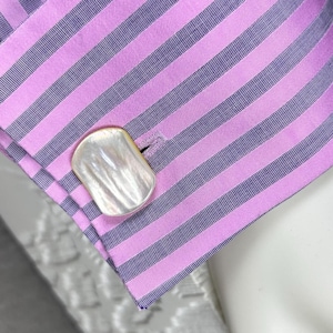 May include: Close-up of a striped pink and gray dress shirt cuff with a mother-of-pearl cufflink. The cuff is buttoned and the stripes are evenly spaced. The cufflink is rectangular with a rounded edge.