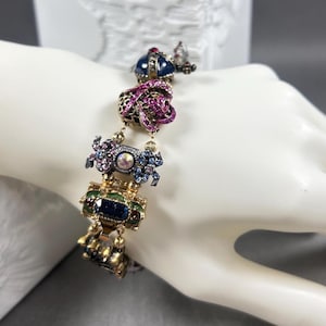 Op de afbeelding: Een goudkleurige armband met sierlijke, met juwelen bezette bedels. De bedels hebben een kroon, een roze lint en andere decoratieve elementen met blauwe, groene en roze edelstenen. De armband wordt getoond op een witte hand.