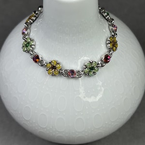 May include: A silver-toned bracelet featuring colorful enameled flower charms. The flowers are in shades of yellow, green, and pink, with small, sparkling accents. The bracelet is displayed on a white, textured surface.