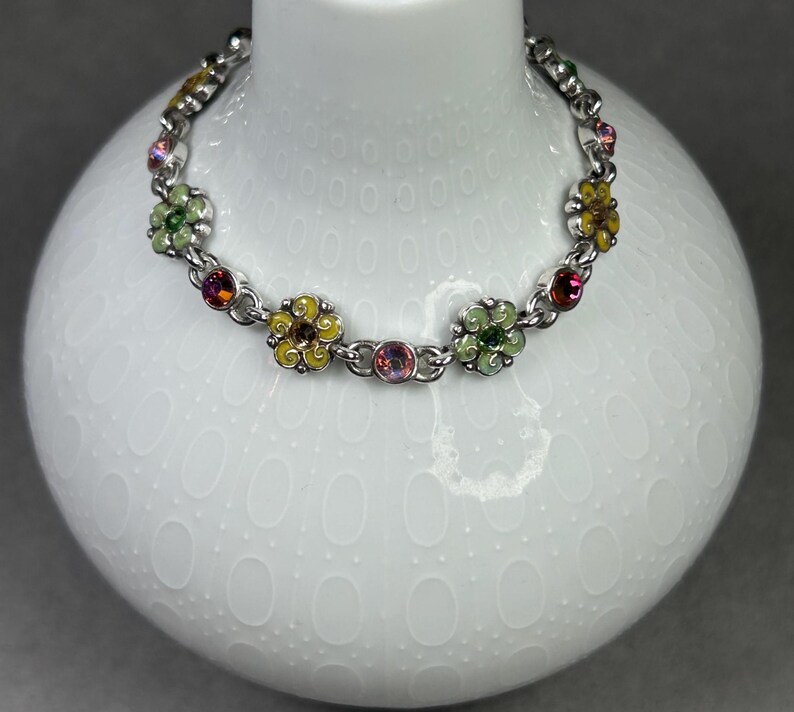 May include: A silver-toned bracelet featuring colorful enameled flower charms. The flowers are in shades of yellow, green, and pink, with small, sparkling accents. The bracelet is displayed on a white, textured surface.