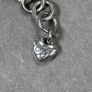May include: Close-up of a silver necklace chain with a heart-shaped charm. The charm has an embossed design. The chain links are circular and connected. The background is a solid grey colour.