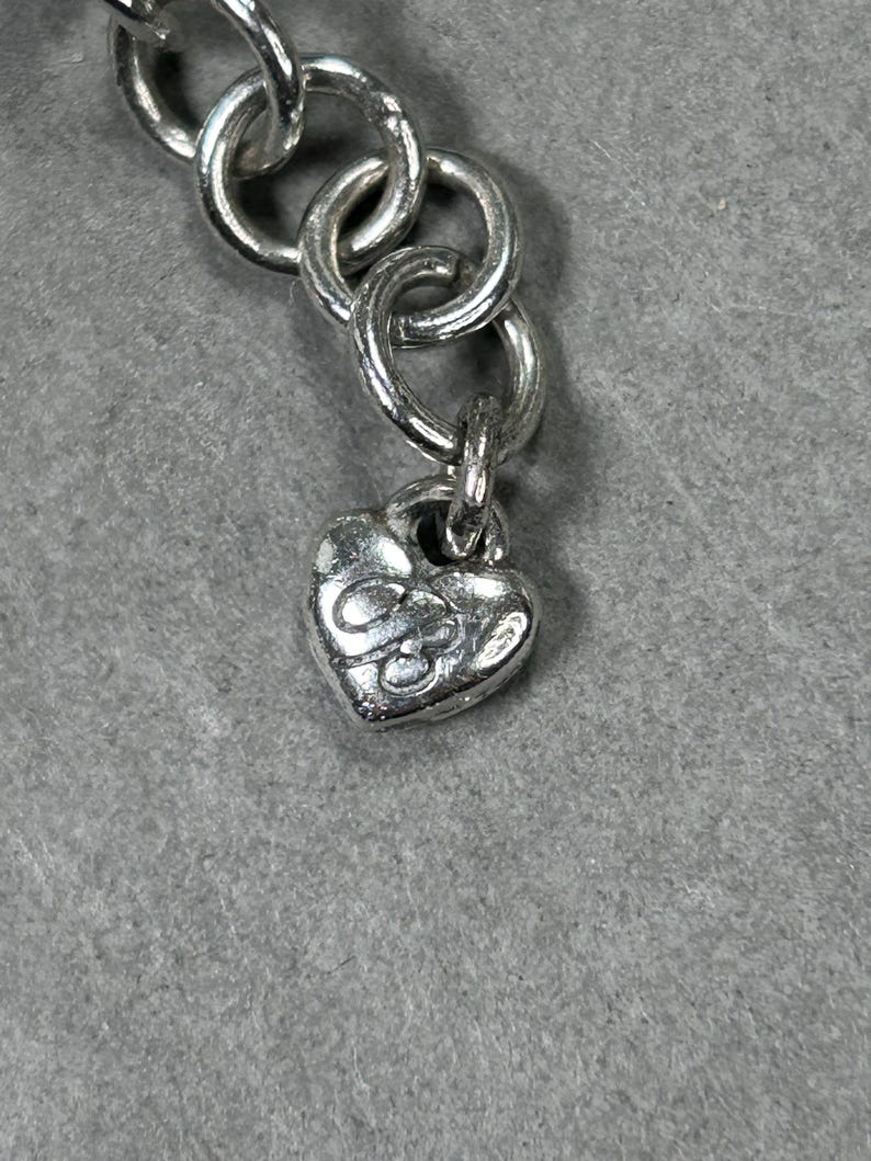 May include: Close-up of a silver necklace chain with a heart-shaped charm. The charm has an embossed design. The chain links are circular and connected. The background is a solid grey colour.