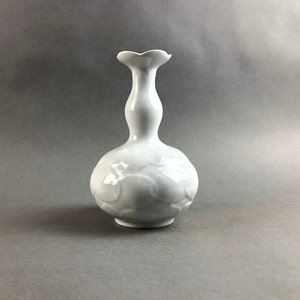 Puede incluir: Un jarrón de cerámica blanca con un cuerpo bulboso y un cuello delgado que se ensancha en un borde festoneado. El jarrón presenta un sutil patrón floral en relieve en la parte inferior. El diseño es elegante y minimalista.