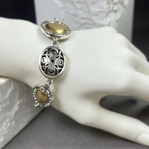 以下が含まれることがあります： ゴールドカラーのアクセントが付いたシルバーカラーのブレスレット。ブレスレットには、楕円形、ハート型、丸型の3つの装飾的なリンクがあります。楕円形のリンクは透かし彫りのデザインです。ブレスレットは白いマネキンの手に表示されています。