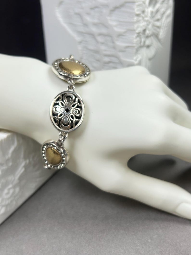 以下が含まれることがあります： ゴールドカラーのアクセントが付いたシルバーカラーのブレスレット。ブレスレットには、楕円形、ハート型、丸型の3つの装飾的なリンクがあります。楕円形のリンクは透かし彫りのデザインです。ブレスレットは白いマネキンの手に表示されています。